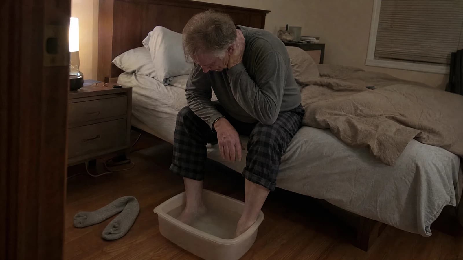 An older man sitting on the edge of his bed at night with his bare feet in a basin of cool water, head down