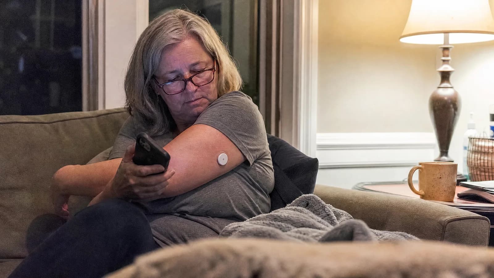A woman in her late fifties on her couch in the evening, checking the glucose sensor on the back of her arm with her phone