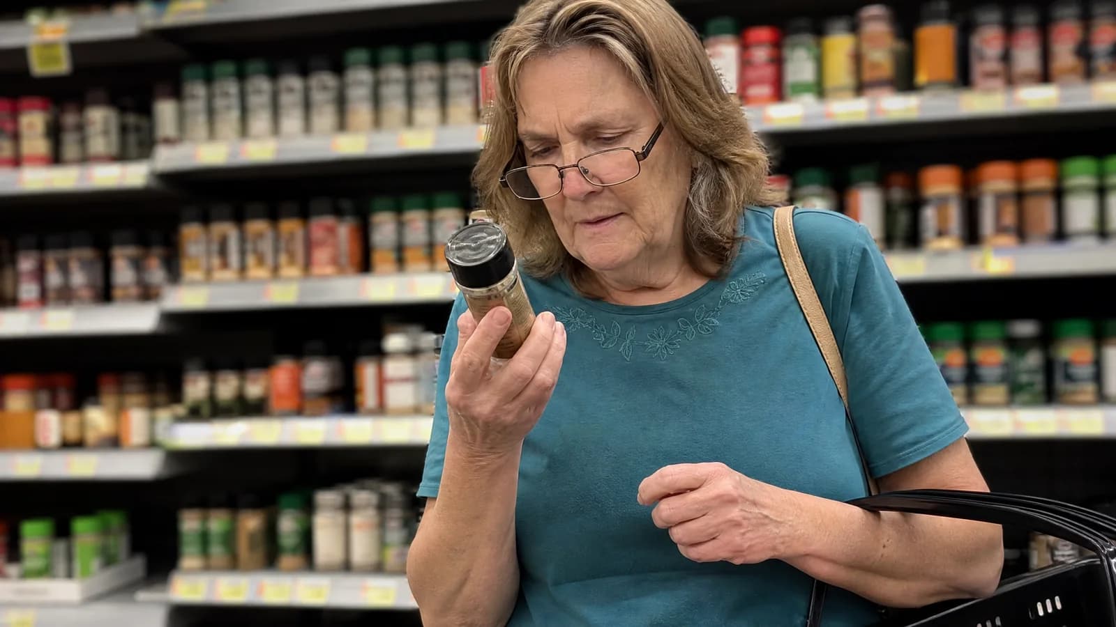 A woman of about 62 in a supermarket spice aisle, reading glasses down her nose, squinting at the small print on a spice jar
