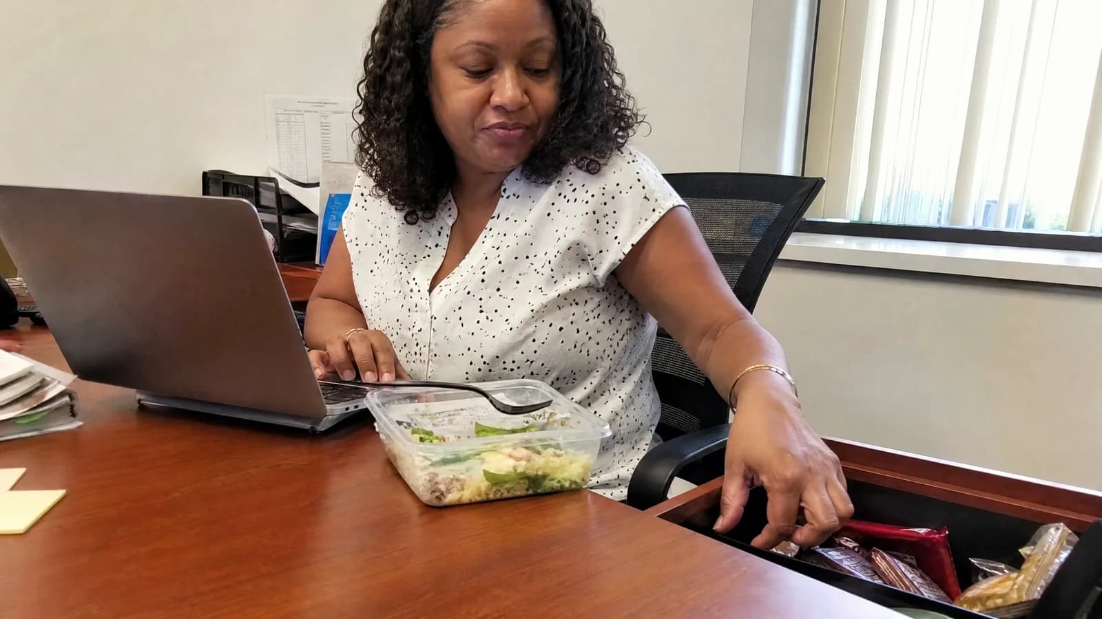 A woman about 48 at her office desk mid-afternoon, reaching into a desk drawer of candy, a half-eaten salad pushed aside