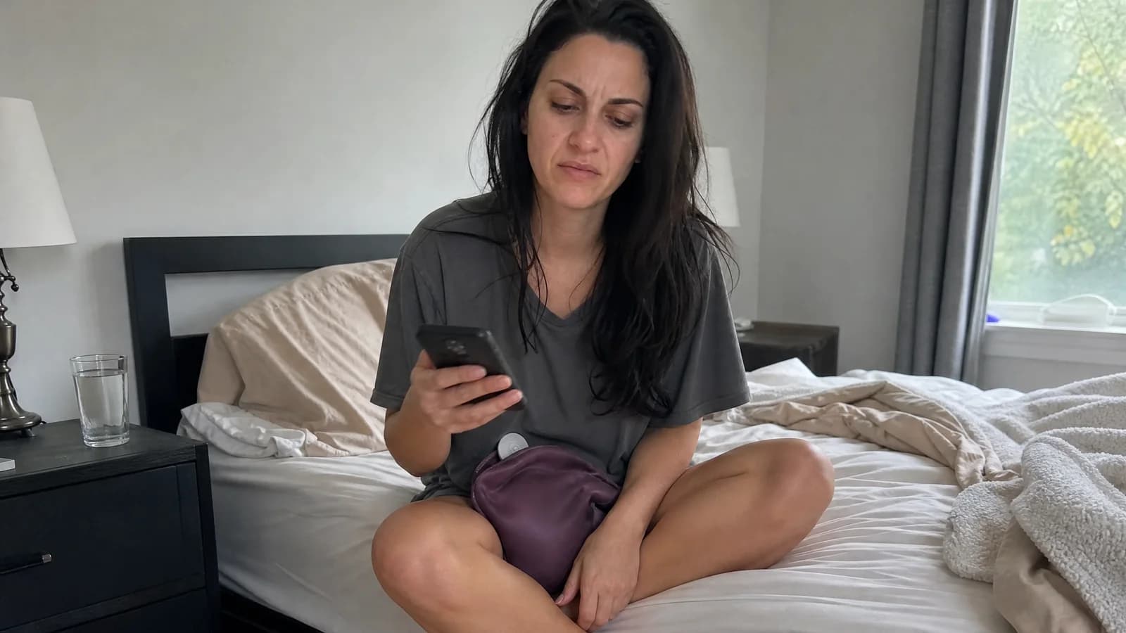 A tired woman in her early thirties sitting cross-legged on her unmade bed in the morning, a hot water bottle on her lap, frowning at her phone