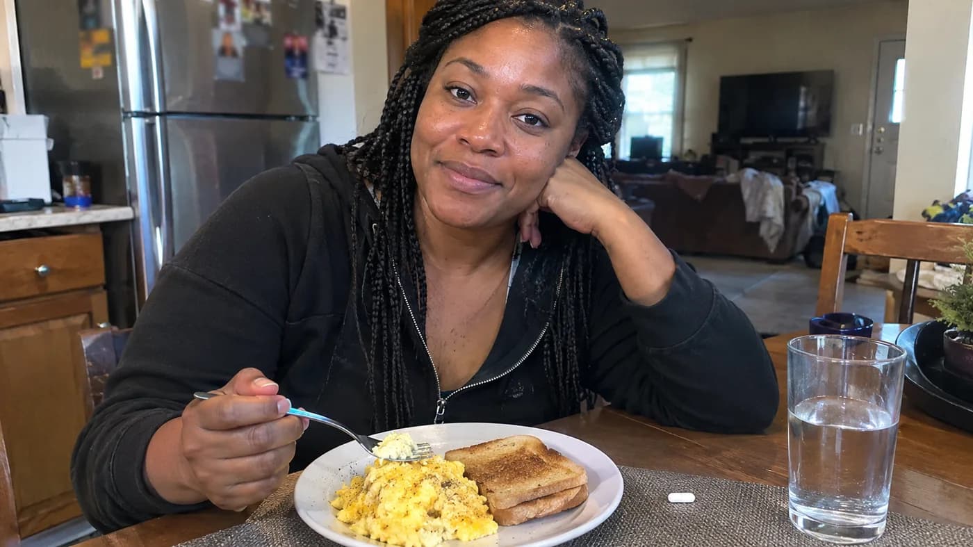 A woman eating breakfast at a kitchen table with a capsule bottle beside her plate