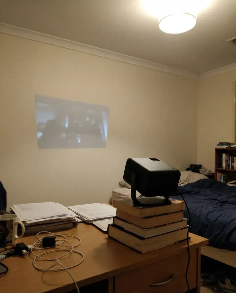 A cheap projector on a pile of books throwing a pale, washed-out picture on a bedroom wall with the ceiling light on
