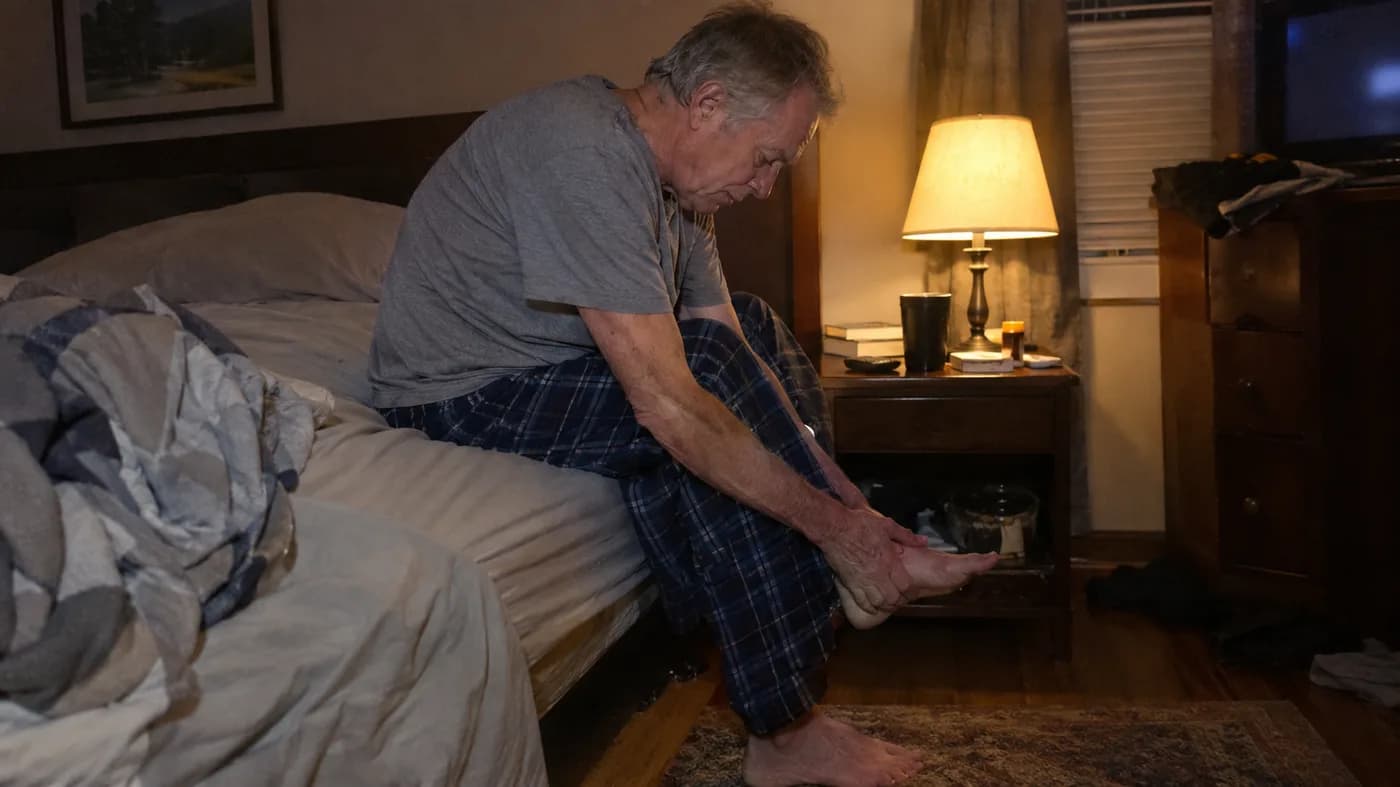 An older man sitting on the edge of his bed at night, rubbing the sole of one bare foot