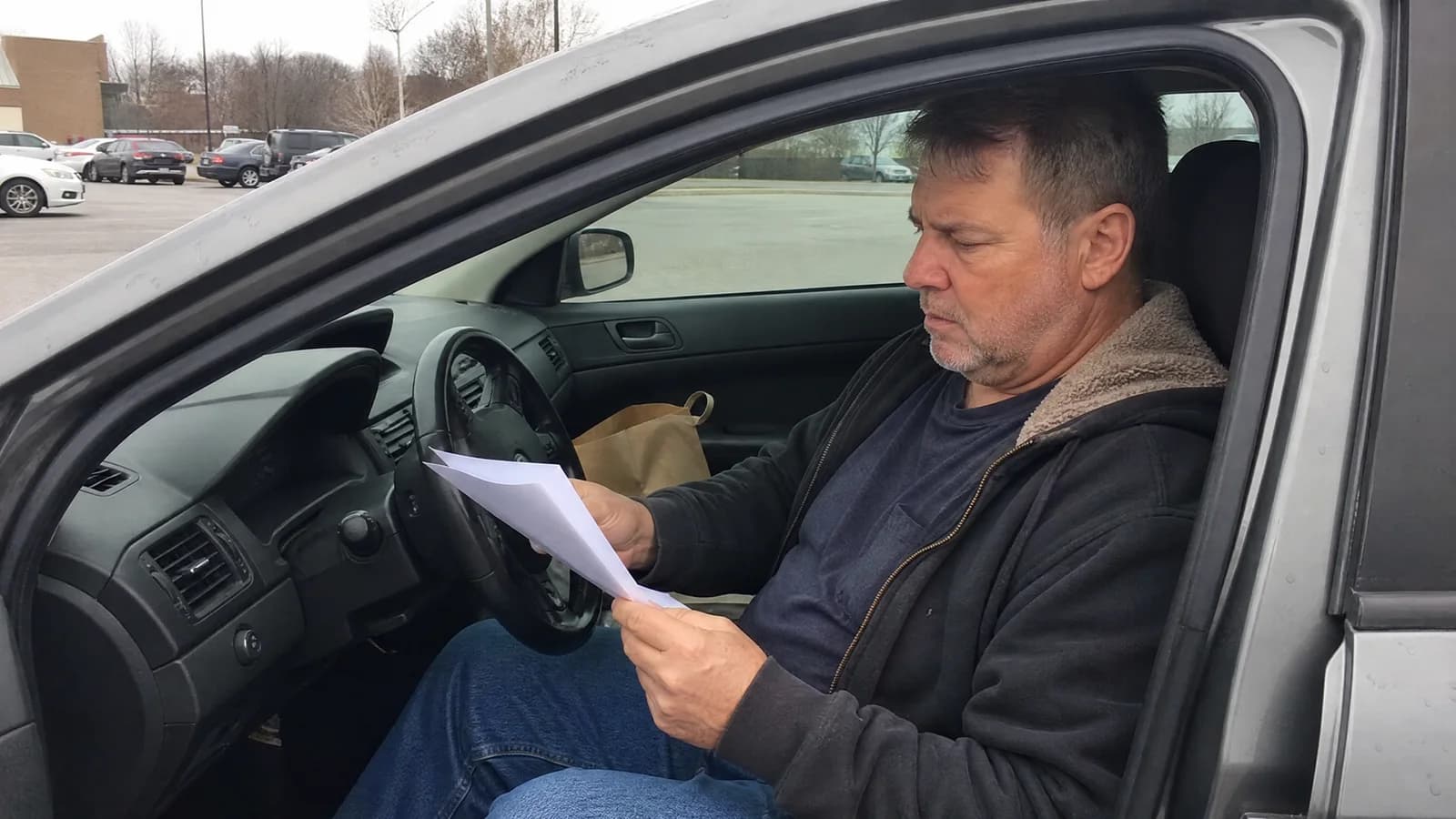 A man in his fifties sitting in his parked car outside a clinic, reading his printed lab results