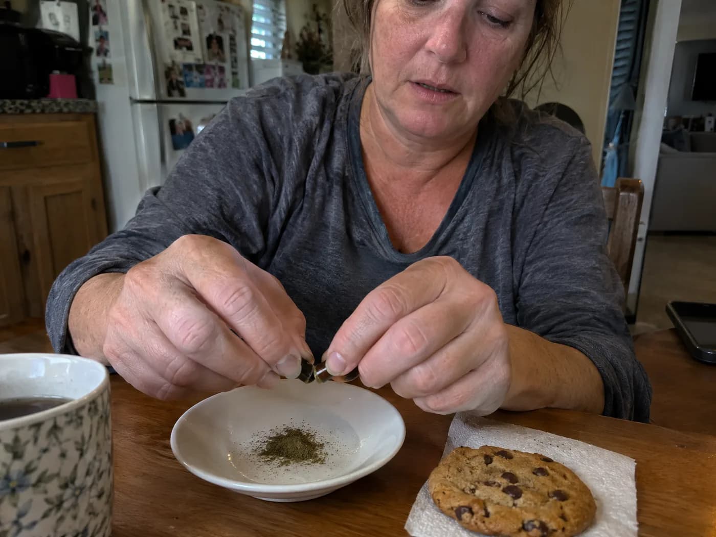 Hands opening a green capsule over a small saucer, a pinch of powder beside a cookie