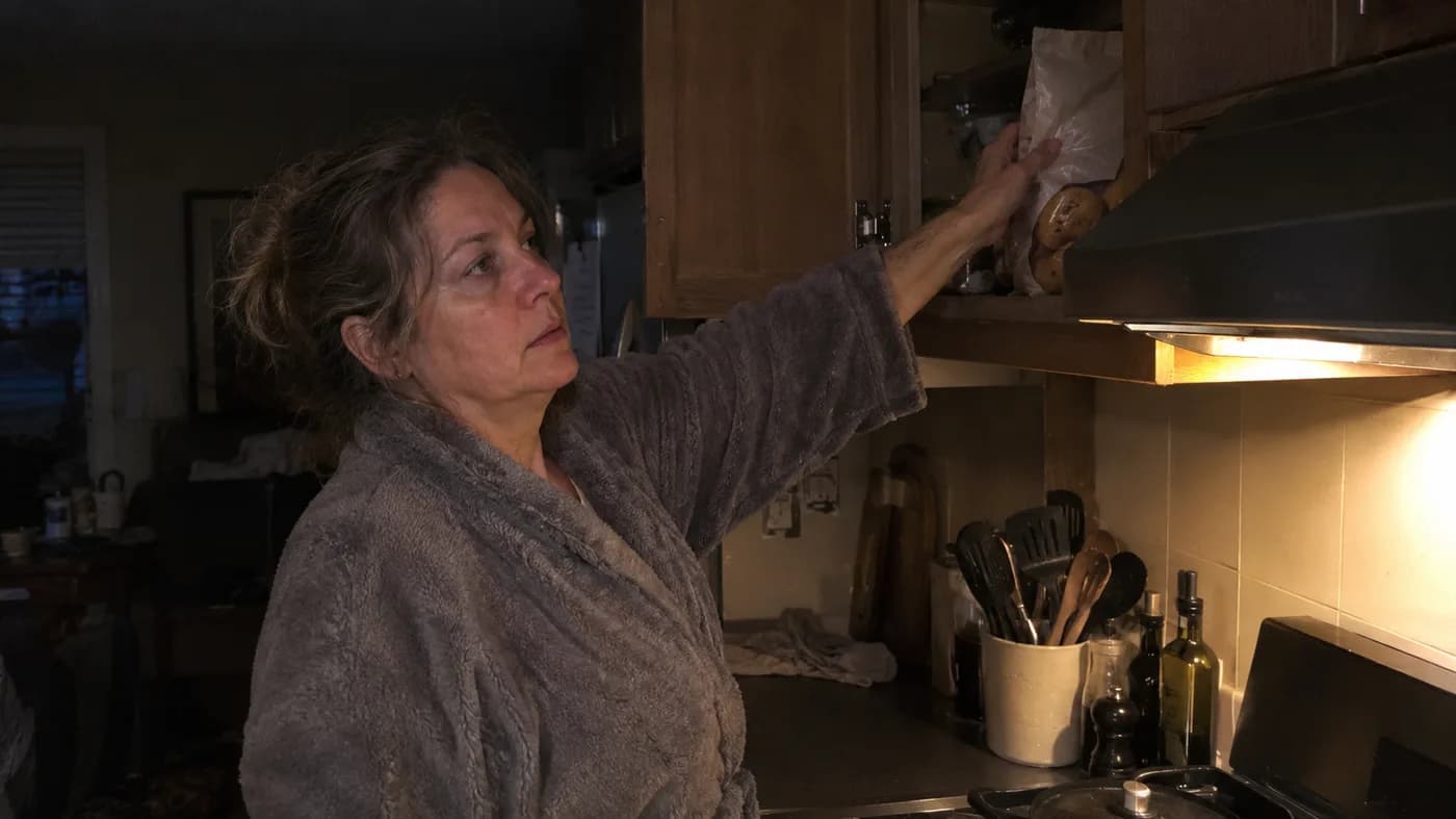 A woman in her fifties standing at an open kitchen cupboard at night, reaching for a bag of cookies